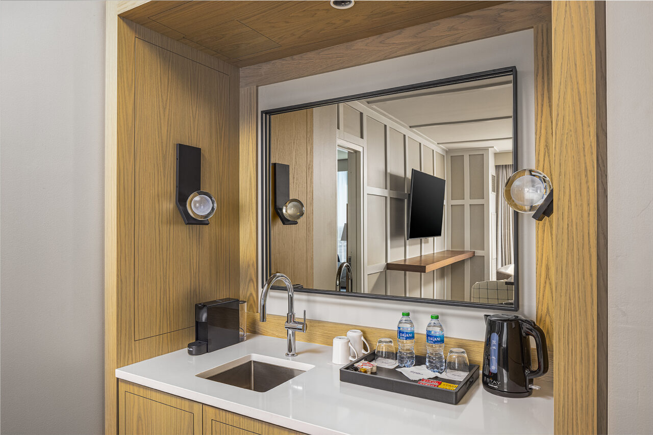 June Prince Suite hotel room kitchenette with a sink, coffee maker, electric kettle, bottled water, and a tray with cups; large mirror reflects TV and modern furnishings in the background.
