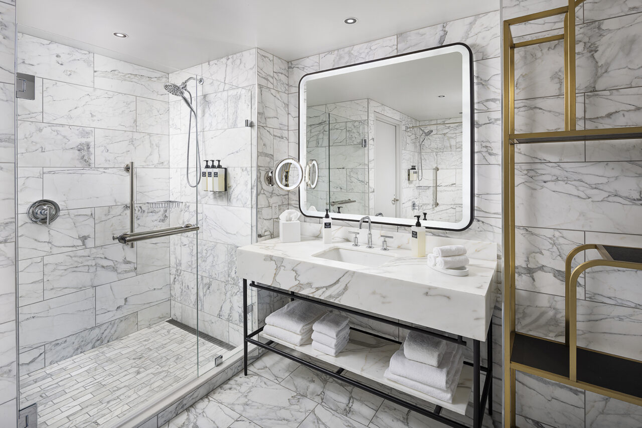 Modern bathroom in the June Prince Suite with marble walls, large lit mirror, double sink, glass shower with bench, and open shelves holding towels and toiletries.