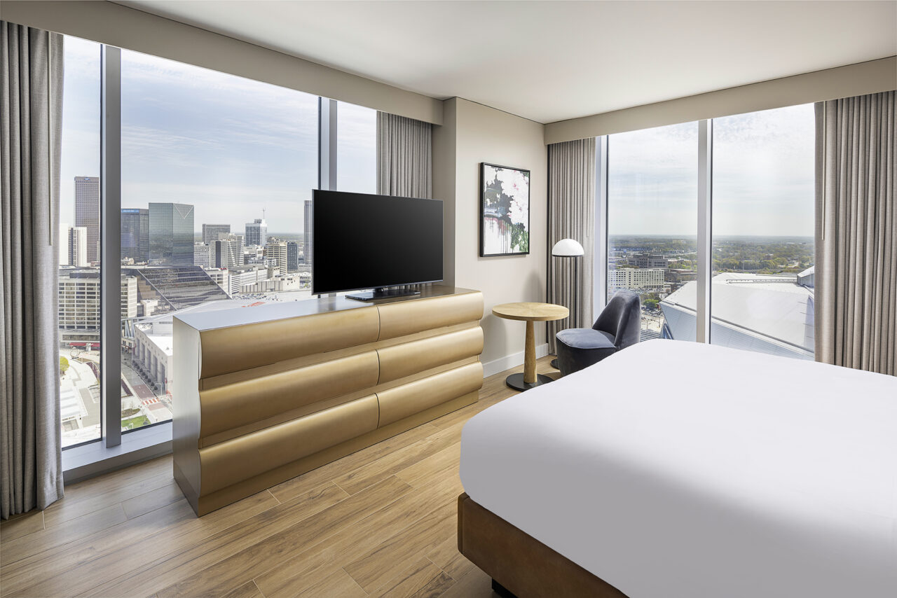 Modern June Prince Suite hotel room with large windows showing a city skyline, a bed, a dresser with a TV, a chair, small table, and a floor lamp.