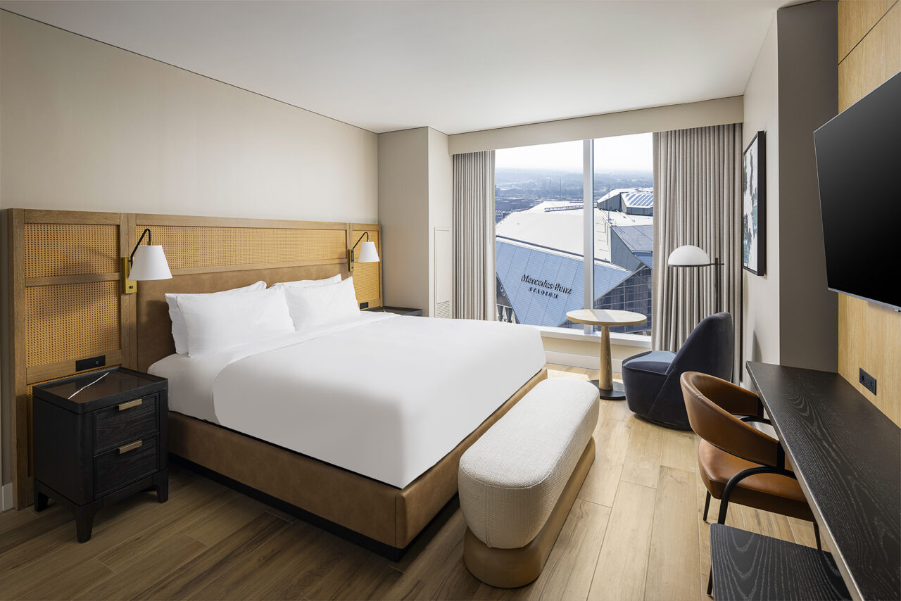 Modern hotel room with a large bed, desk, chairs, TV, and window overlooking a stadium. The June Prince Suite features wood floors and minimalist decor for a stylish and comfortable stay.