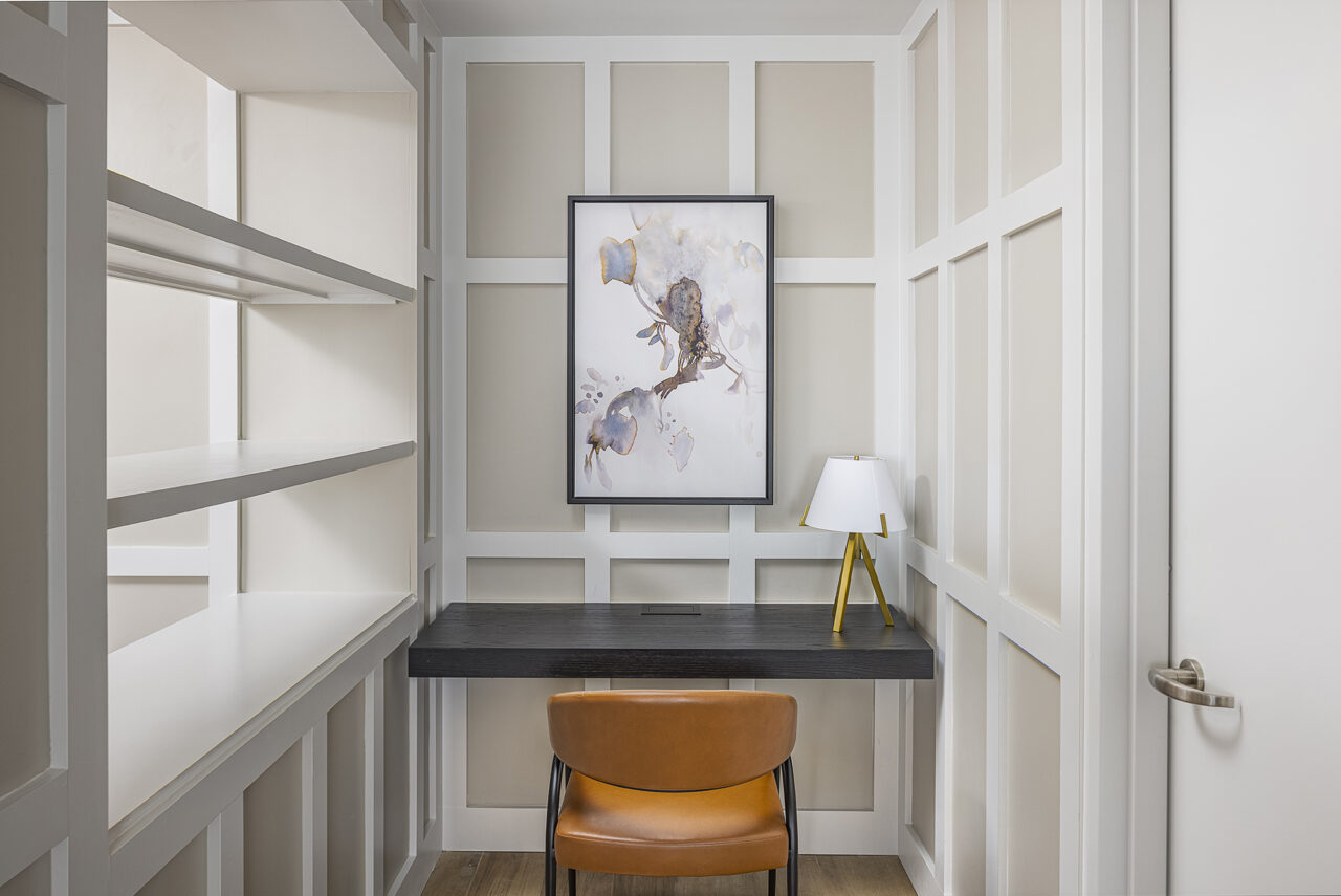 Small home office with white panel walls, black desk, tan leather chair, white lamp with yellow base, and abstract artwork above the desk—perfectly styled for the June Prince Suite.