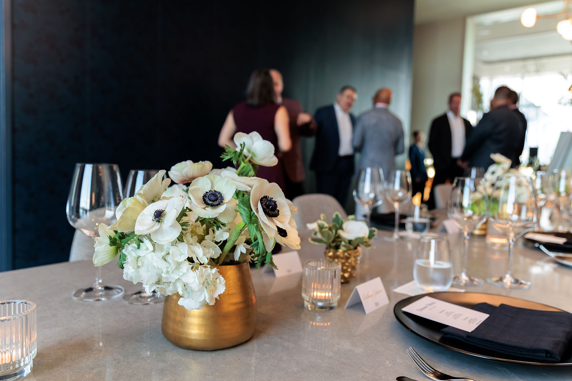 A close-up of a formal private dining table set with white flowers in a gold vase, glassware, and place cards, as guests engage in conversation in the blurred background.