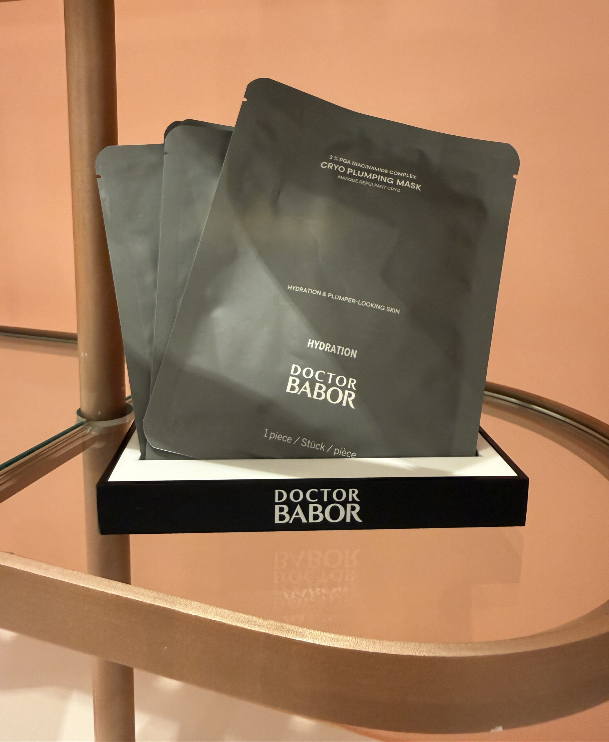 Three Doctor Babor Cryo Plumping Mask packets are displayed upright in a black and white box on a glass shelf with a peach-colored background.