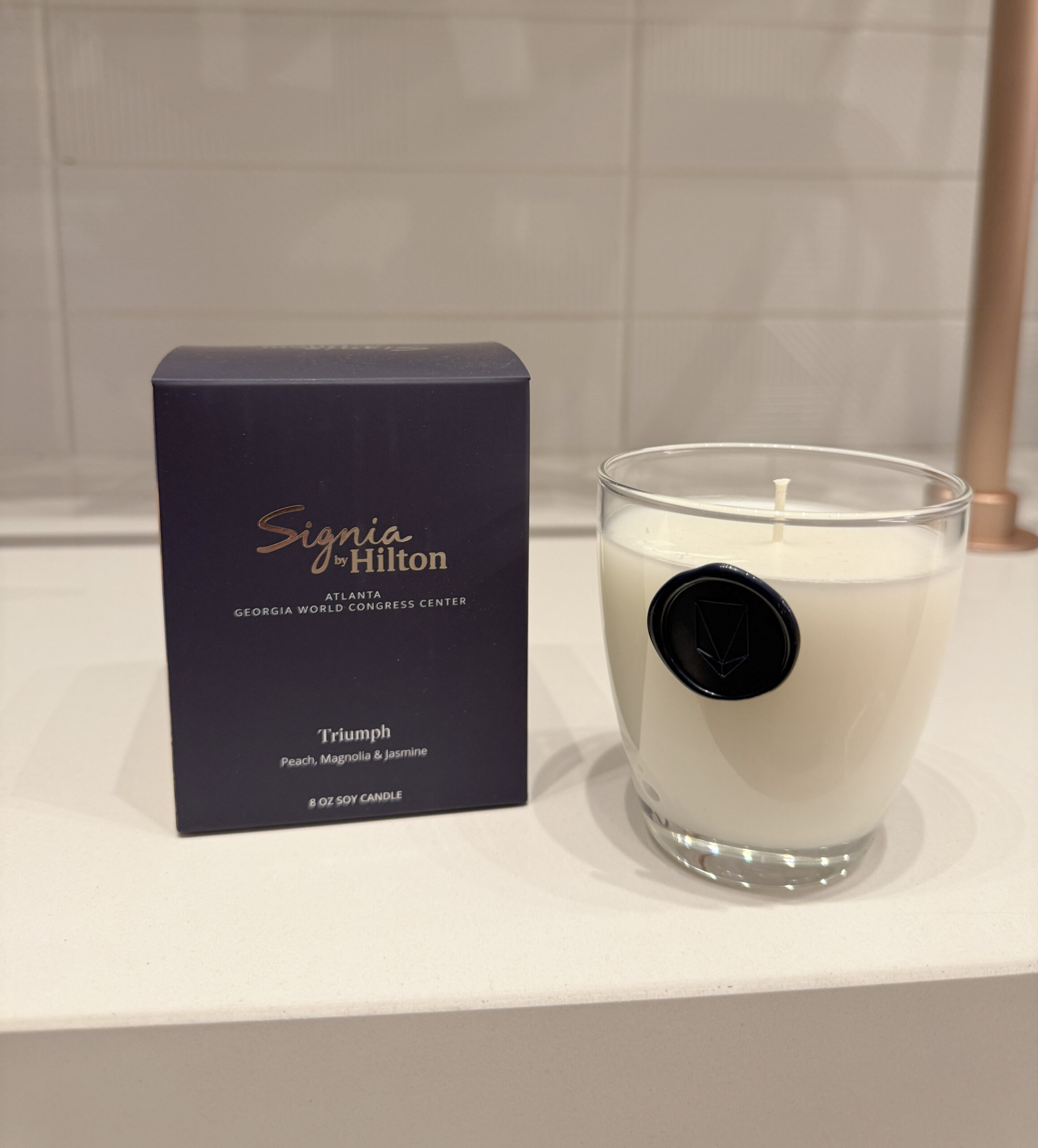 A Signia by Hilton Triumph scented candle sits next to its box, labeled with peach, magnolia, and jasmine scents, on a white countertop with a tiled backsplash.