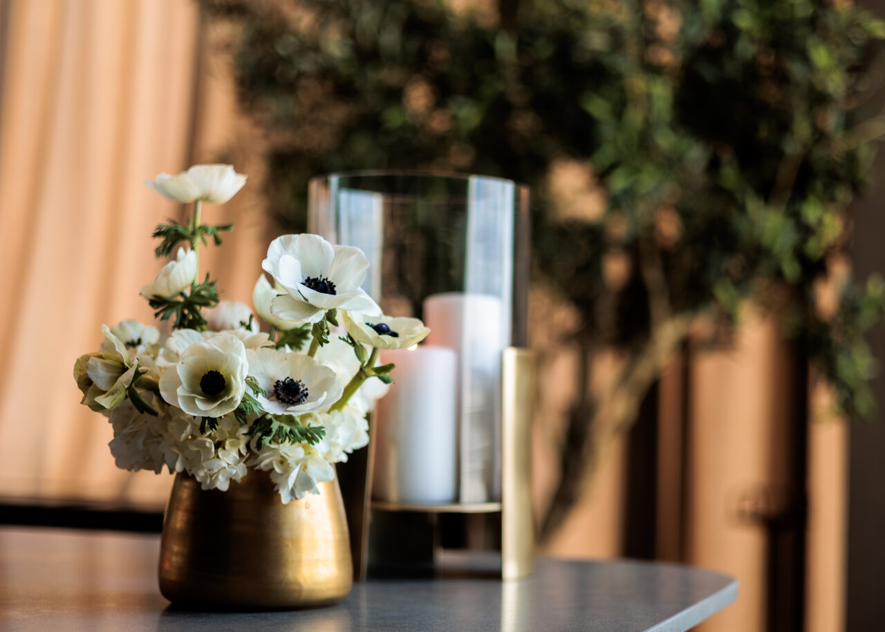 A gold vase with white flowers sits on a table next to a glass hurricane candle holder; a blurred plant is in the background.