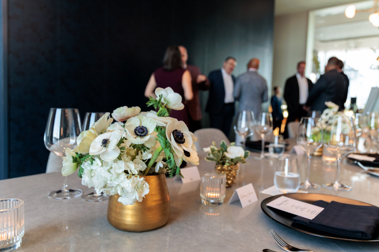 A gold vase with white flowers sits on a set dining table, with several people standing and talking in the blurred background.