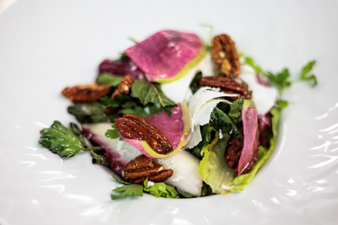 Mixed salad with leafy greens, sliced radishes, pecans, and cheese shavings served on a white plate.