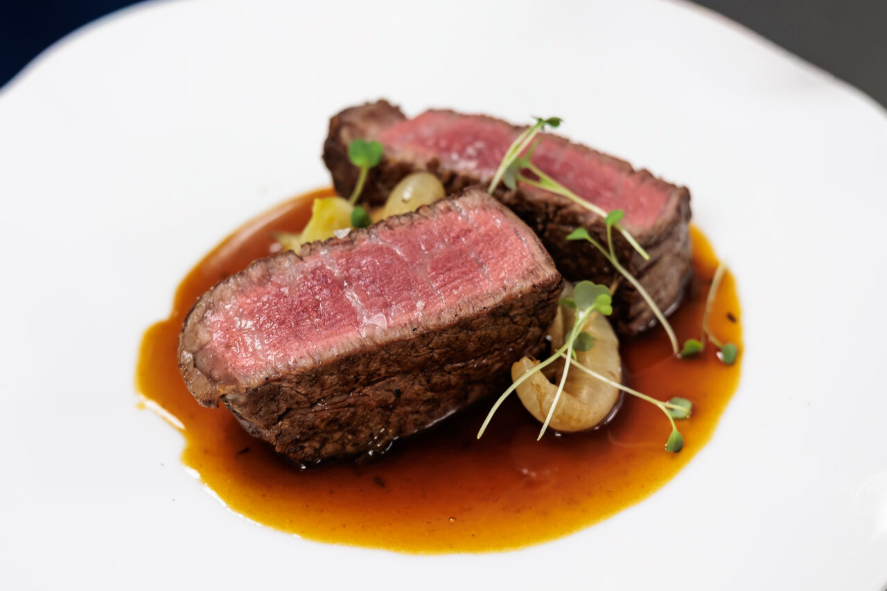 Two slices of medium-rare steak on a white plate with brown sauce, garnished with microgreens and pearl onions.