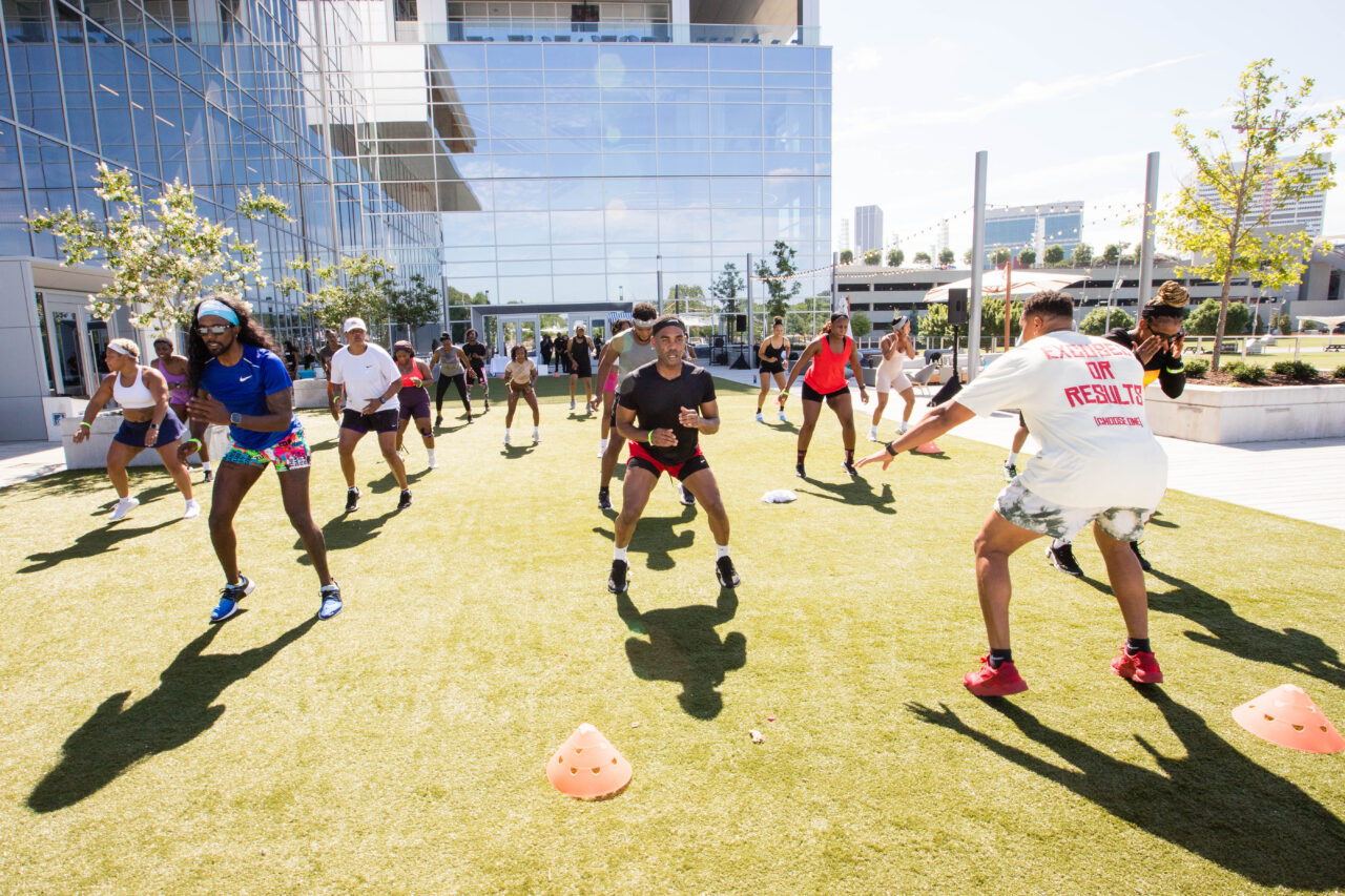 A group of people participates in an outdoor fitness class on a grassy area, with orange cones marking spots and a modern glass building in the background.