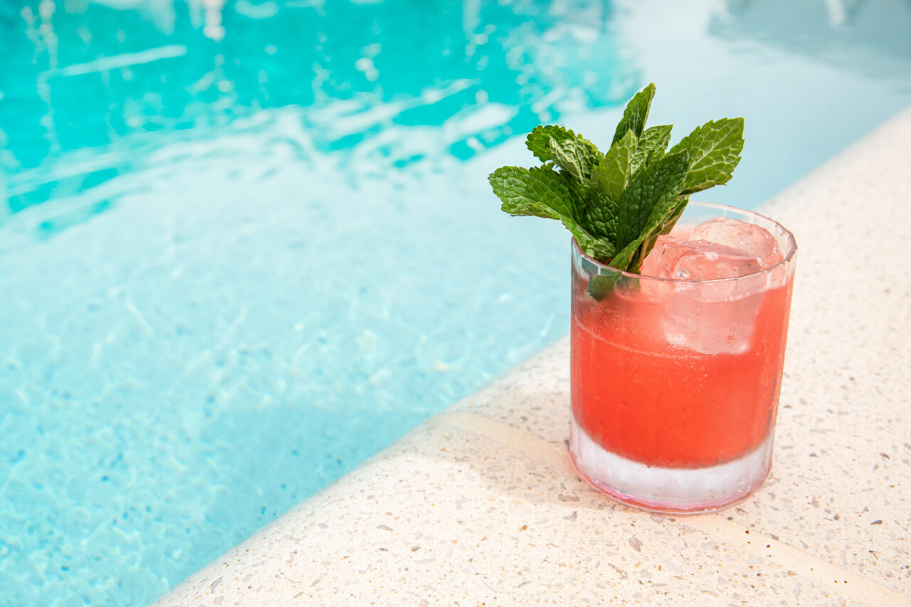A red cocktail with ice and a mint garnish sits on the edge of a pool, next to clear blue water.