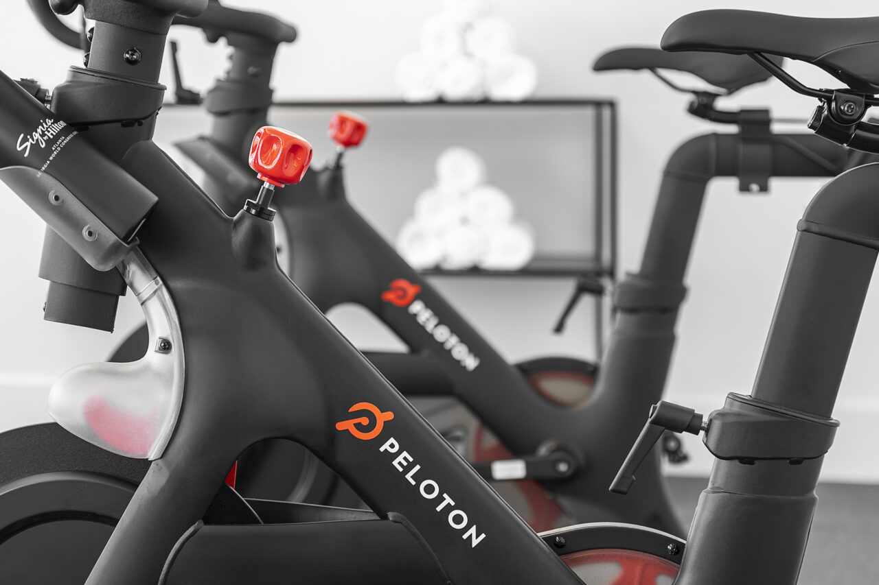 Two Peloton stationary bikes are positioned indoors, with a blurred background featuring a rack holding neatly rolled white towels.