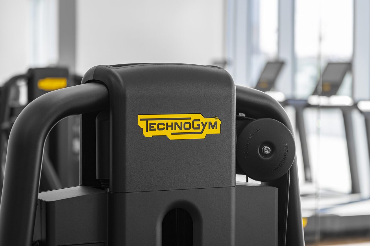 Close-up of a Technogym-branded fitness machine in a gym, with other exercise equipment and large windows visible in the blurred background.