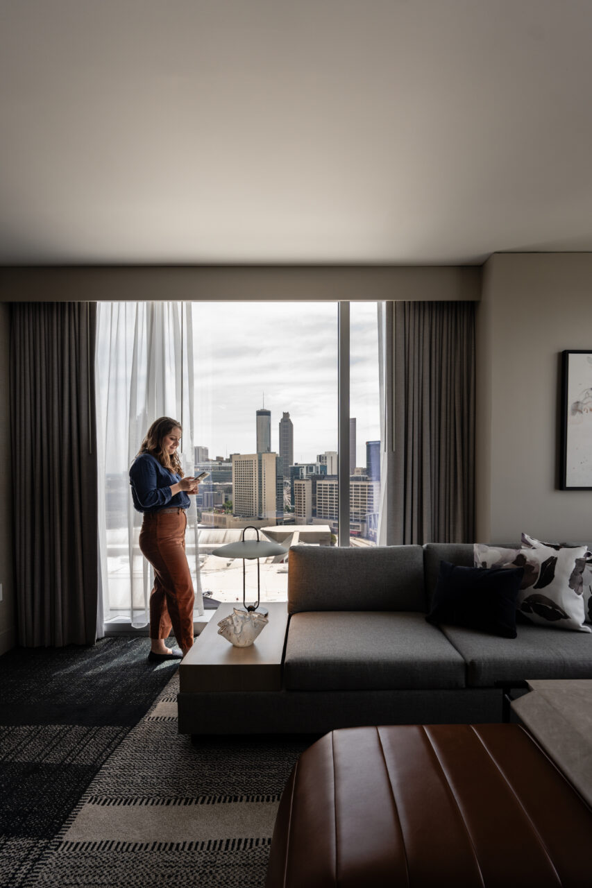 A woman stands by a window in a modern living room, enjoying her suite accommodation during her hotel stay, looking at her phone with the city skyline visible outside.