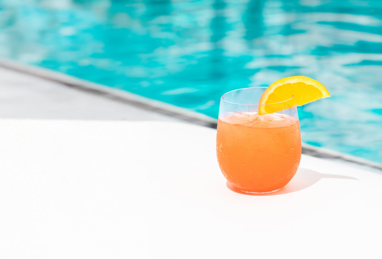 A glass of orange cocktail with ice and an orange slice garnish sits on a white surface next to a swimming pool.