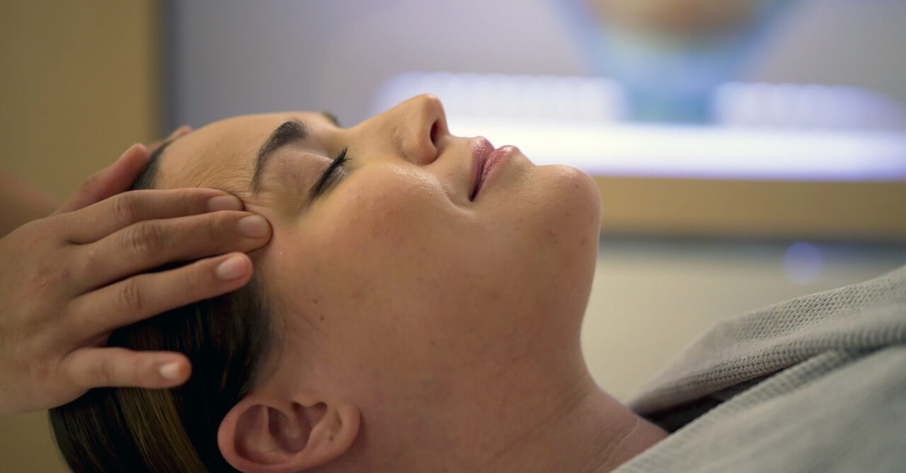 A person lies with eyes closed while someone gently touches their forehead, suggesting relaxation or a facial treatment.