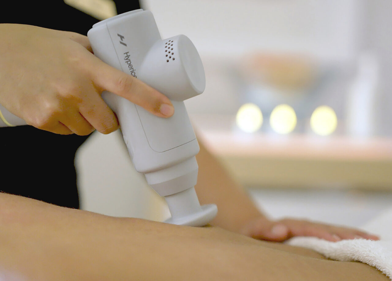 A person uses a white handheld massage device on someone’s arm, with a towel placed nearby on a blurred background.