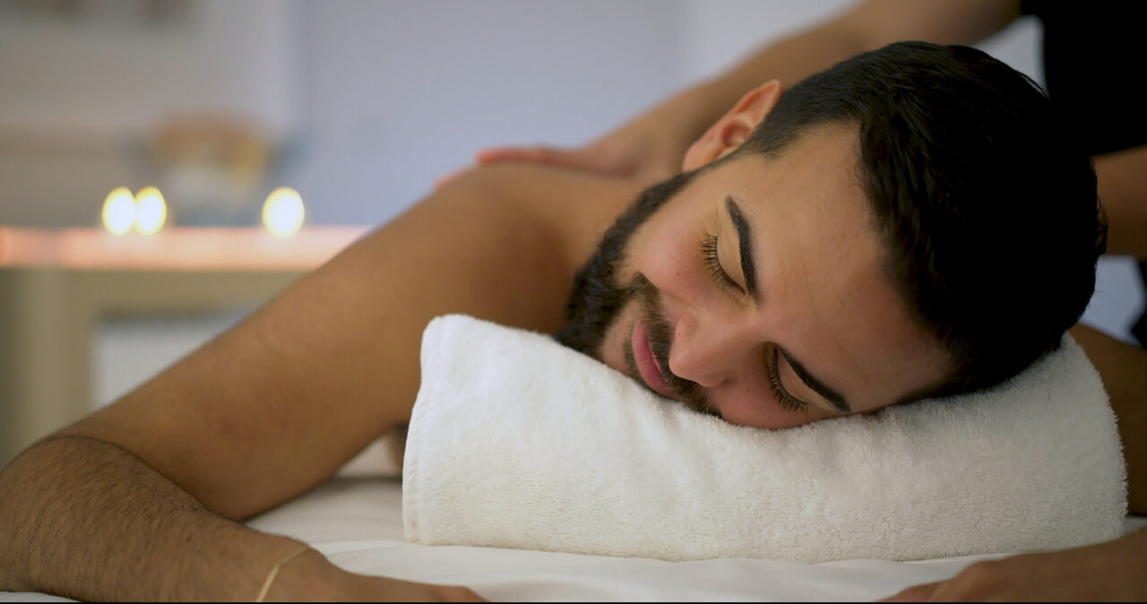 Man with a beard lying face down on a towel, receiving a back massage in a softly lit room, enjoying a soothing treatment designed to detox and renew both body and mind.