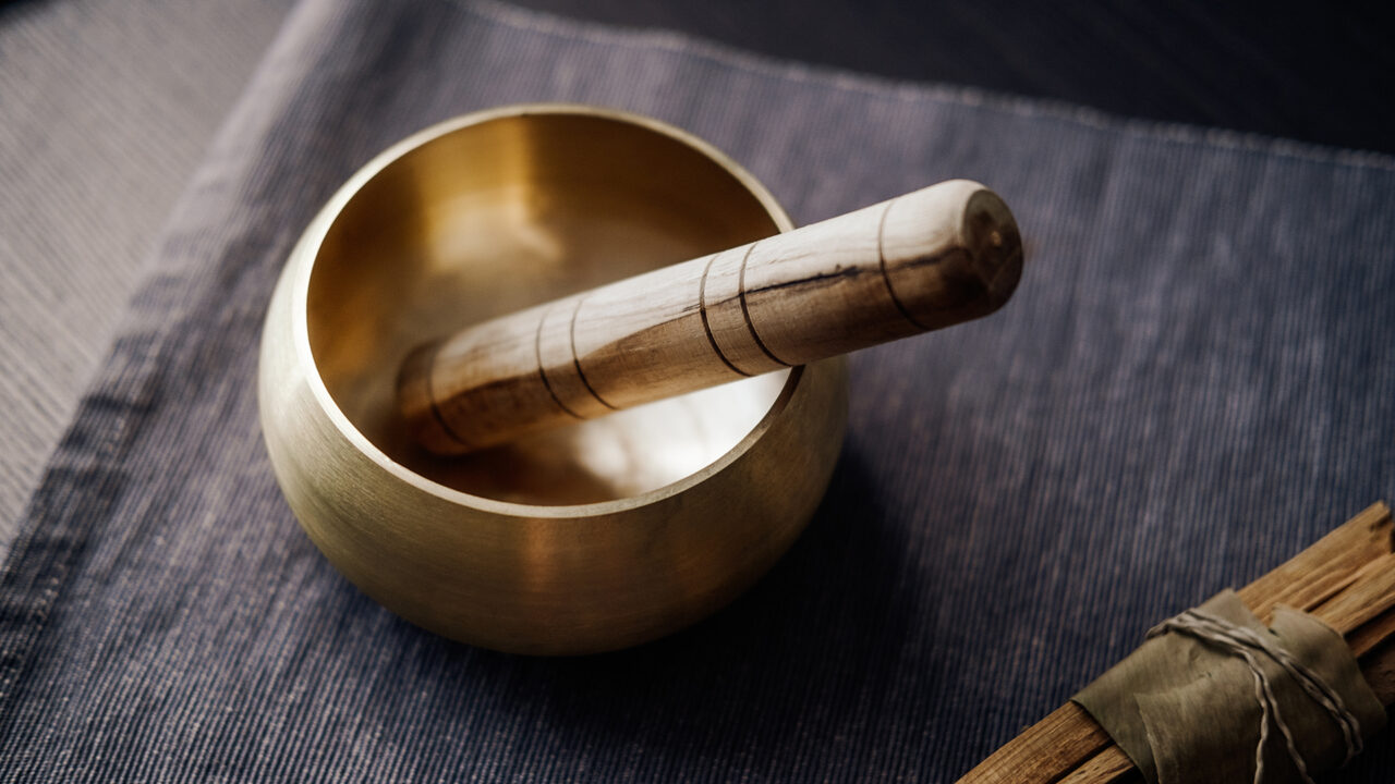 A brass singing bowl with a wooden striker resting inside, placed on a textured dark fabric surface.