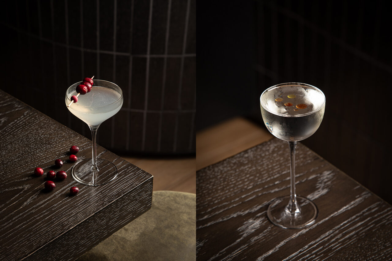 Two cocktails in stemmed coupe glasses sitting on a dark wooden table, one garnished with cranberries and the other with droplets of oil.