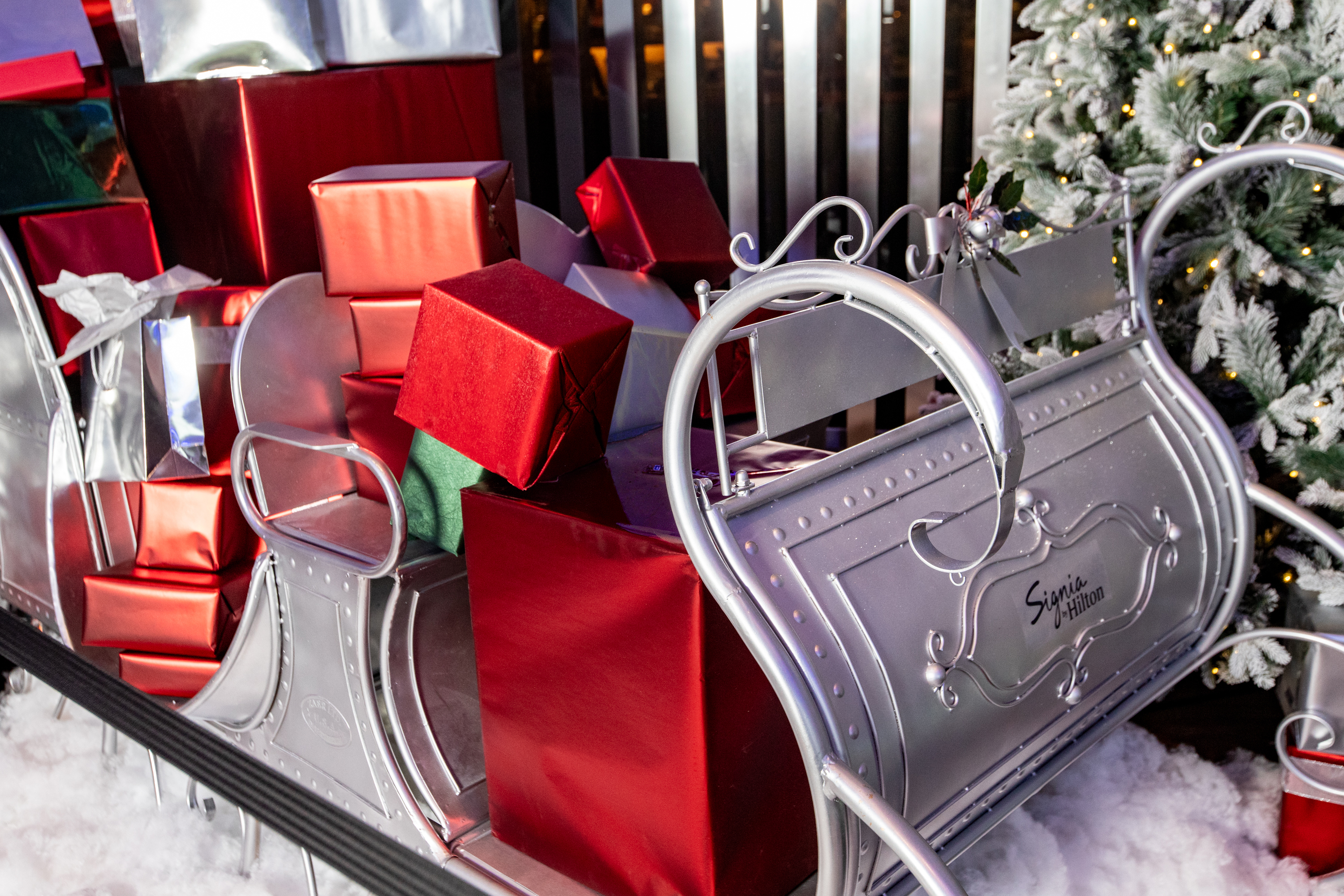Silver Signia sleigh filled with red and silver wrapped gift boxes, positioned next to a Christmas tree on artificial snow.